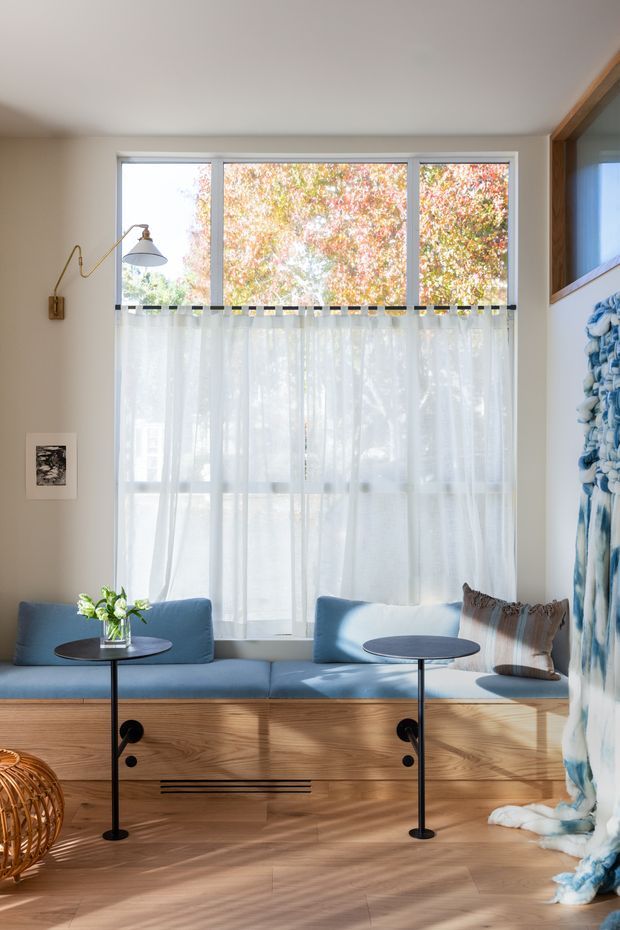 Bright, intimate living room or sunroom with a built-in blue upholstered window seat over warm timber cabinetry, sheer curtains, and large windows overlooking leafy trees. Slim black round side tables, a brass wall lamp, neutral walls, timber flooring, a woven cushion, and a large blue-and-white textile wall hanging create a soft contemporary, natural atmosphere.
