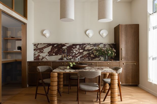 Warm, refined dining and kitchen interior with a compact marble-topped dining table and sculptural cylindrical timber legs, surrounded by upholstered dining chairs. A dark timber kitchen or bar joinery wall includes a dramatic burgundy-and-white veined stone splashback, integrated cabinetry, open shelving, and a tall pantry cabinet. The room has pale neutral walls, timber parquet flooring, rounded pendant lights, decorative wall sconces, and abundant natural light through sheer curtains.