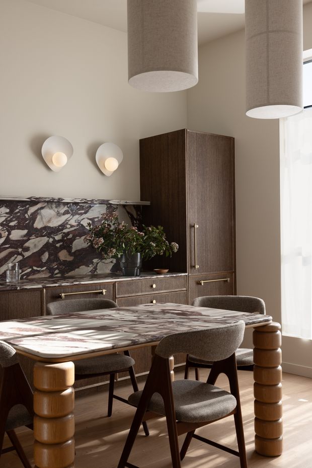A refined dining area with a built-in dark timber sideboard and tall cupboard, dramatic veined stone backsplash and matching dining table, sculptural upholstered dining chairs, rounded timber table legs, and large cylindrical fabric pendant lights. Warm neutral walls, timber flooring, and soft daylight create an elegant, tactile interior.