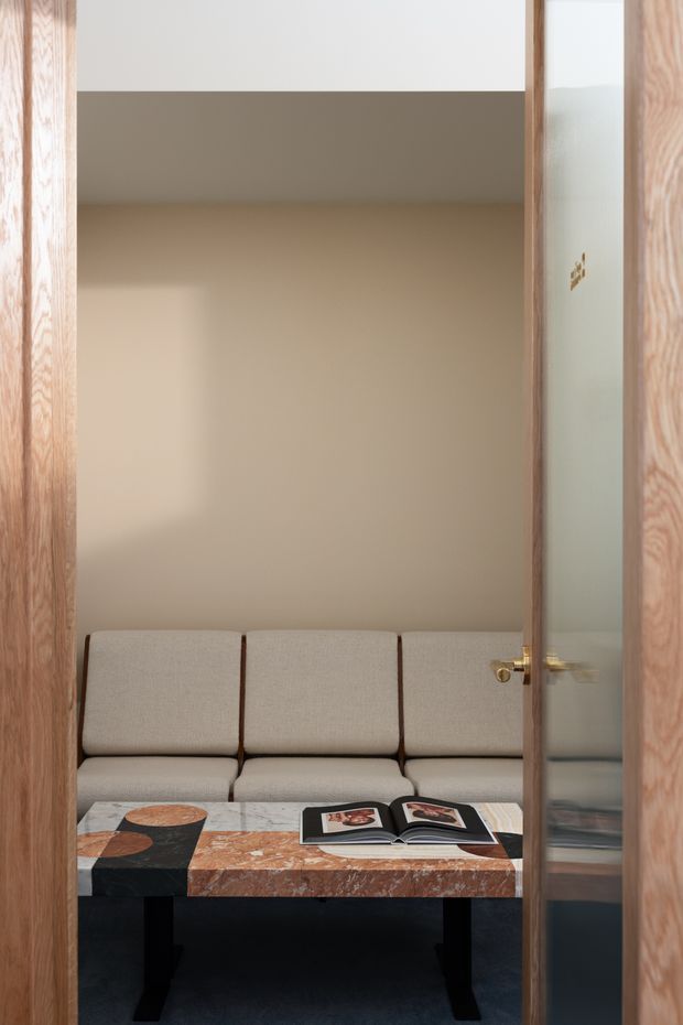 A compact, refined seating nook viewed through an open timber-framed glass door. The room has warm beige walls, a built-in cream upholstered banquette, and a distinctive rectangular marble coffee table with mixed pale, terracotta, and dark stone surfaces on black legs. An open art book rests on the table. The composition is minimalist and warmly lit, with strong timber framing and subtle brass hardware.
