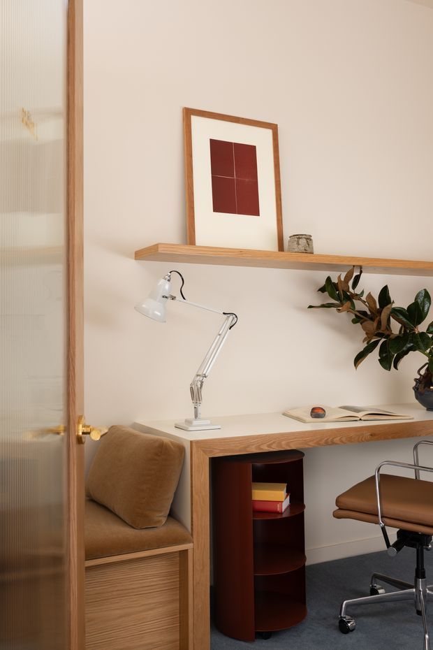 A compact, warmly styled home office viewed through a timber-framed reeded-glass door. The room features a built-in timber desk with open shelving, a white articulated desk lamp, a tan upholstered built-in seat, a wheeled leather office chair, a small dark wood cylindrical storage unit, a potted plant, and framed abstract artwork. The palette is soft cream, natural timber, tan, burgundy, and dark grey, creating a refined mid-century-inspired workspace.