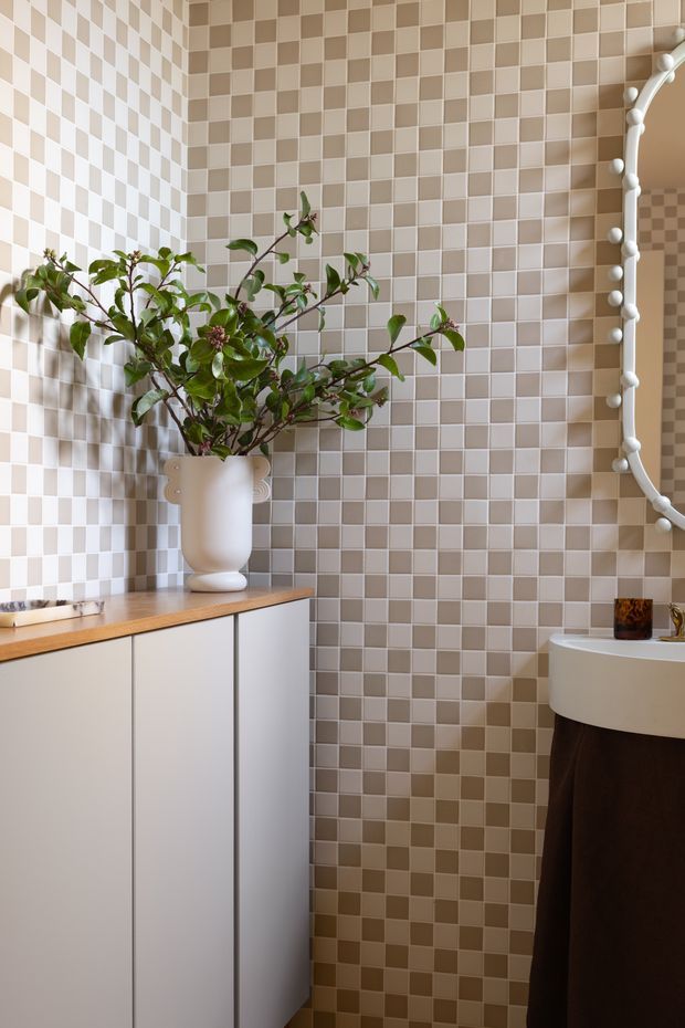 A compact, stylised bathroom corner with walls fully clad in small alternating beige and off-white checkerboard tiles. A minimalist white vanity cabinet with a warm timber benchtop sits beneath a decorative ceramic vase and leafy branches. At right is a partially visible white basin with a dark timber-toned vanity front, alongside a large white-framed mirror edged with spherical bulb lights. The space has a warm, playful, highly patterned interior aesthetic.