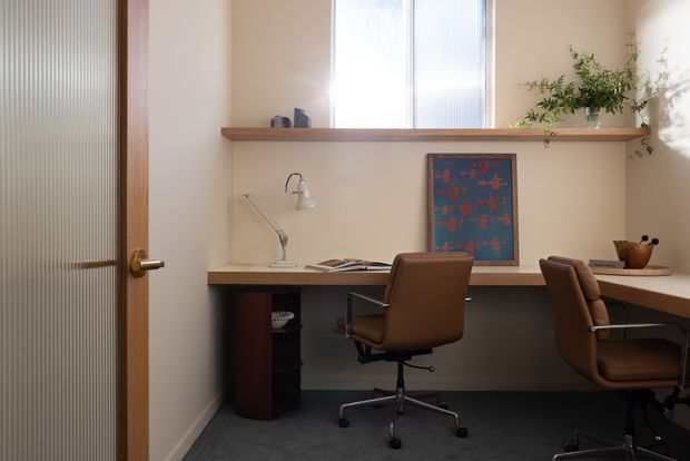 A compact, warmly lit home office with two built-in desk workstations beneath a long timber shelf and frosted vertical-glass windows. The room features a timber-framed ribbed-glass door, neutral plaster walls, dark carpet, brown upholstered office chairs, a desk lamp, artwork, and a potted plant.