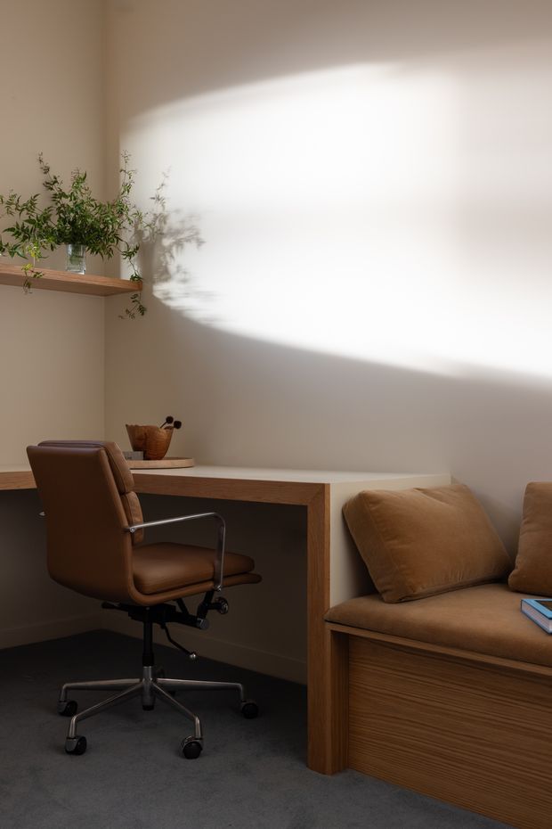 A warm, minimalist home office nook with a built-in timber desk and adjacent built-in window seat or bench upholstered in brown fabric. A brown leather swivel office chair sits at the desk, with a narrow timber wall shelf holding greenery and small decorative objects. Soft sunlight creates a pronounced oval patch of light and plant shadows across the warm off-white wall. The floor is covered in dark grey carpet.