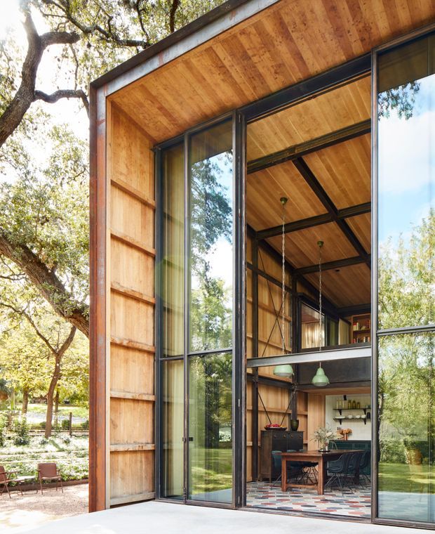 Exterior view of a compact contemporary pavilion-like building set among mature trees, with extensive floor-to-ceiling glazing and dark metal frames. The facade and soffit are clad in warm timber, while the exposed interior reveals a dining and kitchen area, timber ceiling, dark structural framing, shelving, pendant lights, and patterned flooring. The glazed corner strongly connects the interior to the landscaped garden.