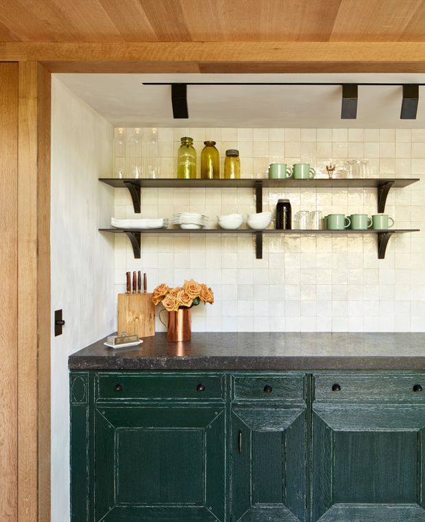 A compact kitchen or kitchenette nook framed in warm timber, with dark green shaker-style cabinetry, a dark stone benchtop, cream square-tile splashback, and open black timber shelves displaying glassware, ceramics, and bottles. The space has a crafted, rustic character with visible timber ceiling and surround.