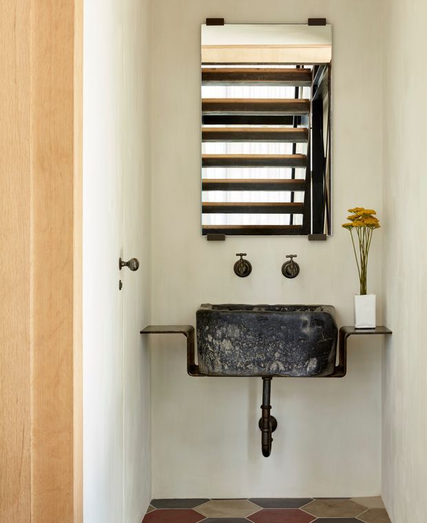 A compact powder room with a wall-mounted dark stone basin set into a dark metal console, exposed plumbing, twin wall taps, and a tall rectangular mirror with timber-toned horizontal detailing. Pale walls, a timber door, and geometric multicoloured floor tiles create a refined rustic character.