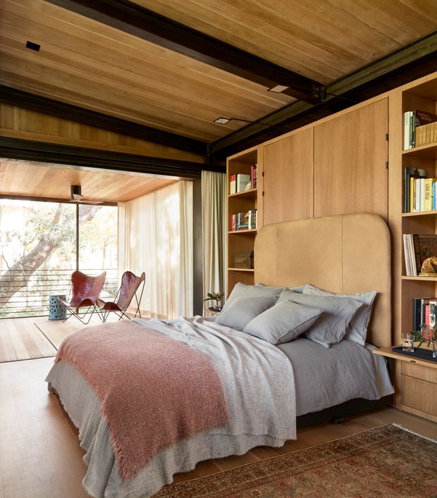 Warm, refined bedroom with a built-in timber bedhead and full-height timber cabinetry incorporating open bookshelves. The room features exposed dark structural beams, a timber-lined ceiling, pale timber flooring, soft grey bedding, and a muted terracotta throw. A glazed sitting area with two lounge chairs is visible beyond the bedroom, bringing in abundant natural light and views of trees.