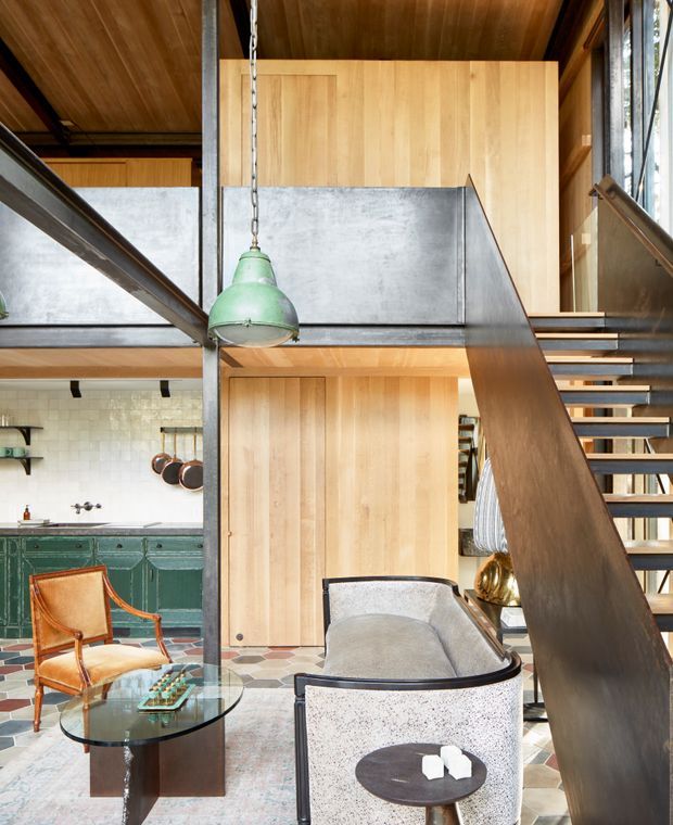 A tall, open-plan interior combining a living area, kitchen, and open stairway with mezzanine-level circulation. The space features exposed dark steel structural elements and stair balustrades, warm timber wall and ceiling lining, a green vintage-style pendant light, green cabinetry, patterned tiled flooring, and a mix of leather and upholstered lounge furniture. Large glazing at right brings in natural light.