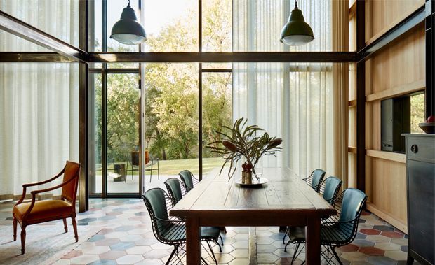 A light-filled dining room with a long solid timber dining table and wire-frame chairs, surrounded by expansive floor-to-ceiling glazing with sheer curtains. Warm timber wall panelling and built-in cabinetry frame the room, while a patterned geometric tile floor and oversized pendant lights add visual interest. Garden greenery is visible through the glass.