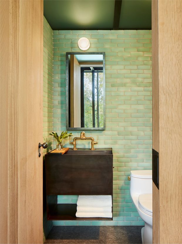 Compact bathroom viewed through a timber door, featuring mint-green glazed brick wall tiles, a dark floating vanity with copper-toned exposed plumbing and basin tapware, a rectangular mirror, white toilet, dark floor tiles, and a dark green ceiling.