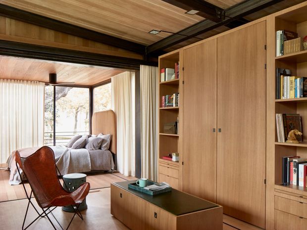 Warm, refined interior showing a timber-lined living room with extensive built-in shelving and concealed cabinetry. A low timber storage unit with a dark top sits in the foreground, while a leather sling chair and coffee table face a sofa in the adjoining glazed sitting area. Floor-to-ceiling curtains and large windows bring in natural light, and exposed dark ceiling framing adds an industrial architectural note.