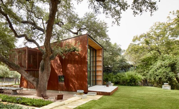 A compact contemporary pavilion clad in rust-red weathered metal or timber-like panels, set within a landscaped garden beneath mature spreading trees. The building has a simple pitched roof, large glazed openings, and a projecting deck or terrace with outdoor seating. A broad lawn and dense greenery surround the structure.