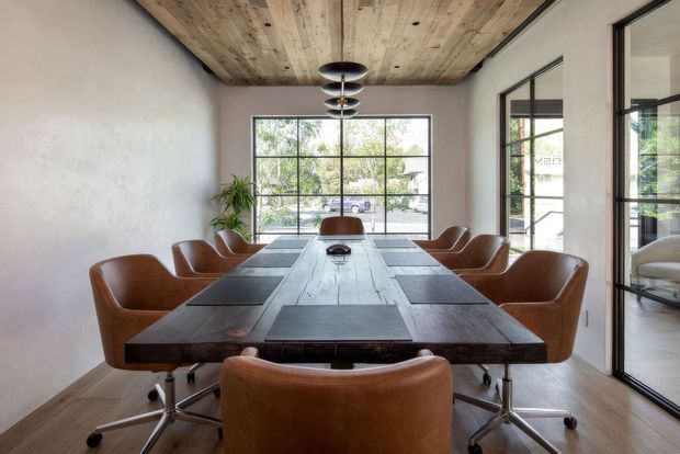 A refined dining room with a long dark timber dining table surrounded by tan leather swivel chairs. The room features a rustic exposed timber ceiling, pale textured walls, timber flooring, black-framed floor-to-ceiling glazing, and a sculptural modern pendant light. Indoor planting and greenery visible outside add a natural element.
