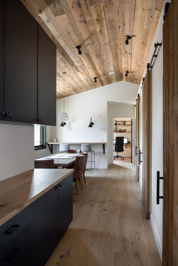 A warm, narrow open-plan interior combining a kitchen and dining area, with a home office visible through a doorway. The space features matte dark cabinetry, light timber flooring and a striking reclaimed-looking timber plank ceiling with exposed knots and varied boards. A compact dining table with upholstered chairs sits beneath pendant lighting, while black wall sconces, black hardware and slim shelving add contrast. The white walls and timber finishes create a refined, contemporary rustic atmosphere.