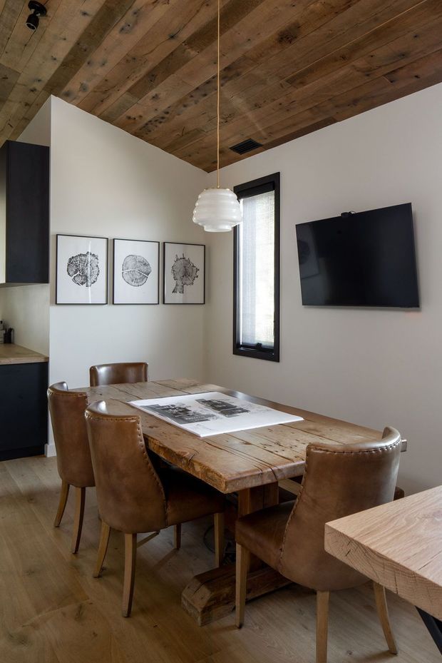 A warm, rustic dining area with a substantial reclaimed timber dining table and matching timber ceiling boards. Four brown leather dining chairs surround the table, while a black-framed narrow window, wall-mounted television, framed botanical-style artworks, and partial dark kitchen cabinetry are visible. A sculptural white pendant light hangs over the table.