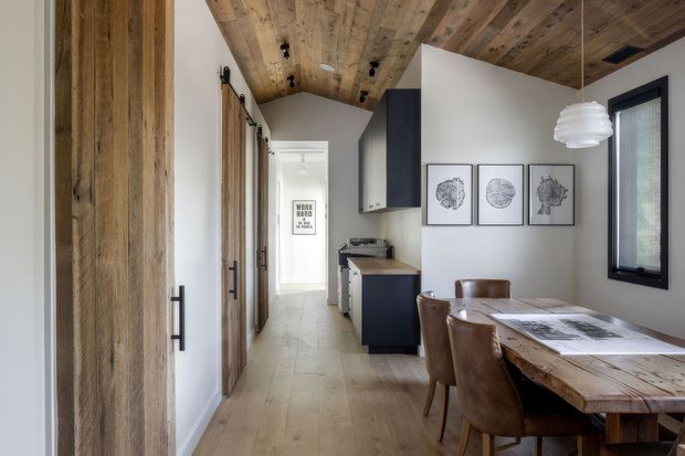A narrow, open-plan dining and kitchen area with a long rustic timber dining table and leather chairs in the foreground. The space features white walls, pale timber flooring, extensive reclaimed timber wall and ceiling cladding, dark kitchen cabinetry, framed botanical artwork, and a black-framed window. A corridor with timber doors extends beyond the kitchen.