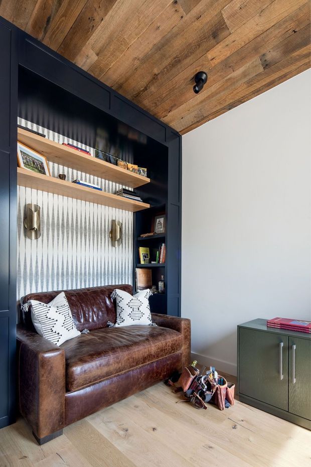 A refined compact study or reading nook with a deep brown leather loveseat set against dark built-in shelving and cabinetry. The joinery includes open timber shelves, decorative lighting, books and framed objects, with a patterned textured wall backing. Reclaimed-looking timber boards line the ceiling, while pale timber flooring and a muted green cabinet create contrast. A small collection of art and stationery items rests on the floor beside the sofa.