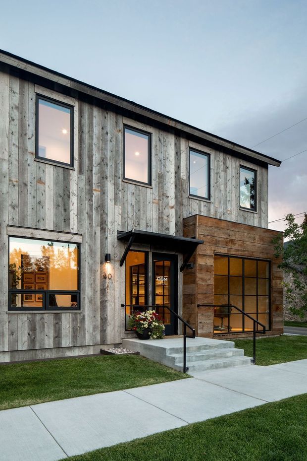 Exterior view of a contemporary two-storey building with weathered vertical timber cladding, dark-framed rectangular windows, a projecting timber-clad entry volume, and a glazed ground-floor frontage. Concrete steps and a sidewalk lead to the entrance, with neatly maintained lawn and a small planted pot at the doorway.