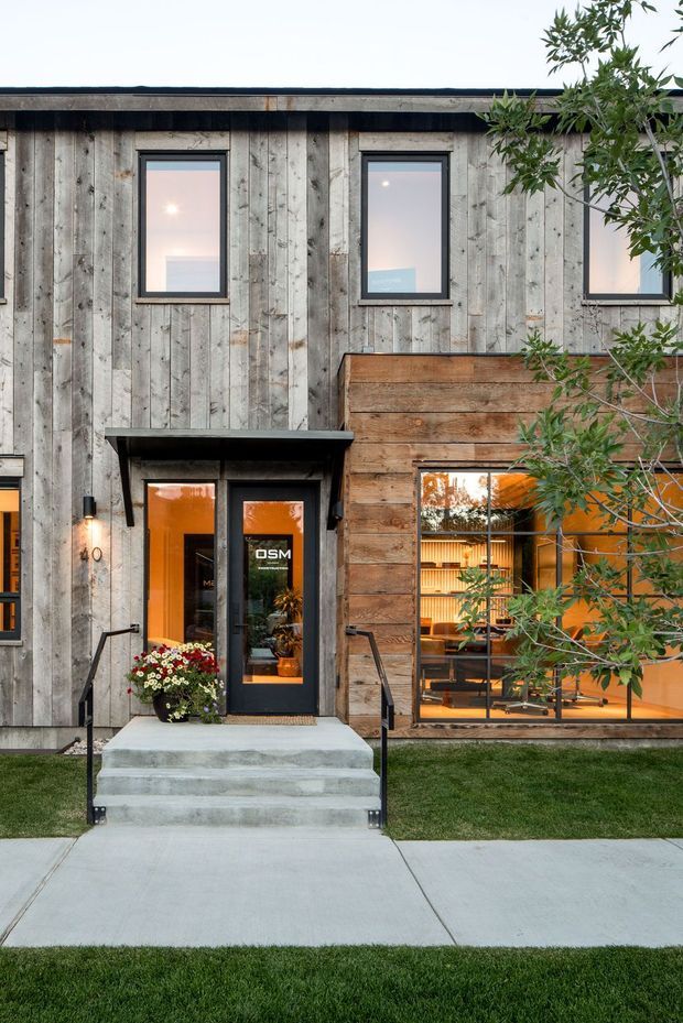 Street-facing facade of a contemporary low-rise building with weathered vertical timber cladding, a contrasting horizontal timber section, dark-framed windows, and a glazed entrance beneath a shallow black metal canopy. Concrete steps and path lead to the doorway, with warm interior lighting visible through the ground-floor glazing and landscaped planting in the foreground.