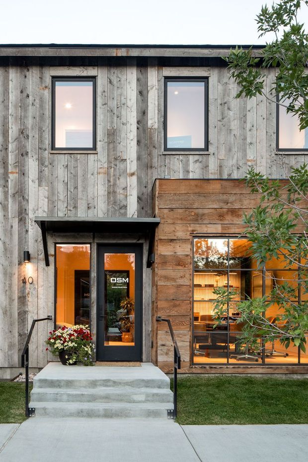 Street-facing exterior of a compact contemporary commercial building, featuring weathered vertical timber cladding, a contrasting horizontal timber-clad projecting section, dark-framed windows and glazed entry doors, concrete steps, black metal handrails, and warm interior lighting visible through the glazing. Landscaping and a small lawn frame the entrance.