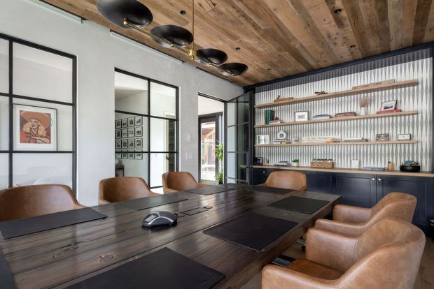 A sophisticated dining and meeting room with a long dark timber table surrounded by cognac leather armchairs. The room features a built-in dark cabinetry and open shelving wall displaying books and decorative objects, black-framed glazed partitions and doors, a pale plaster wall, and a striking reclaimed timber plank ceiling with black pendant lighting.