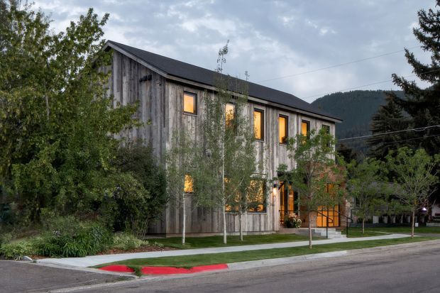 Exterior view of a contemporary, elongated multi-storey building clad in weathered vertical timber boards, with a simple dark pitched roof and regularly spaced warm-lit windows. The building sits within a landscaped suburban or mountain-town streetscape, with mature trees, a sidewalk, and wooded hills visible behind it.
