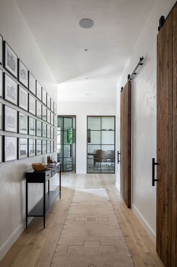 A long, bright residential hallway with pale timber flooring and white textured walls. One wall is arranged as a dense gallery of framed monochrome artwork above a slim black console, while the opposite wall features large rustic timber sliding barn-style doors with black metal hardware. Glazed internal doors at the end reveal a light-filled office or living area. A neutral patterned runner extends through the corridor, creating a refined, warm transitional interior.