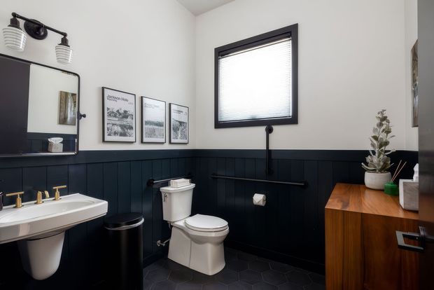 A compact, dark-accented bathroom featuring white fixtures, a wall-mounted basin with brass tapware, a toilet, black hexagonal floor tiles, and dark navy or charcoal vertical wall panelling. A timber vanity cabinet, framed artwork, mirror, and potted plant add warmth and contrast. The window has a black frame and blind, and the room has a clean, contemporary character.