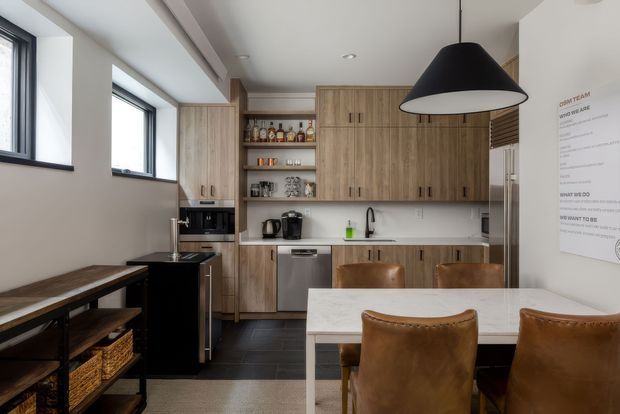 A contemporary open-plan kitchen and dining area with warm wood cabinetry, dark tiled flooring, a pale stone benchtop, integrated shelving, and black-framed windows. A large black pendant light hangs above a white dining table with tan leather chairs. A compact secondary sink and bench are visible along the left wall, while the main kitchen includes a dishwasher, cooktop, and concealed storage.