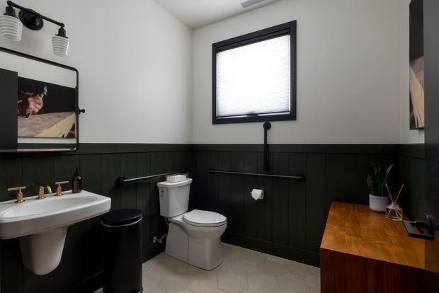 A compact bathroom with white walls, dark green-black vertical wall panelling, a black-framed window with blind, white pedestal basin and toilet, black tapware and accessories, and a warm timber vanity or counter. The room has a strong monochrome palette with natural wood accents and practical traditional detailing.