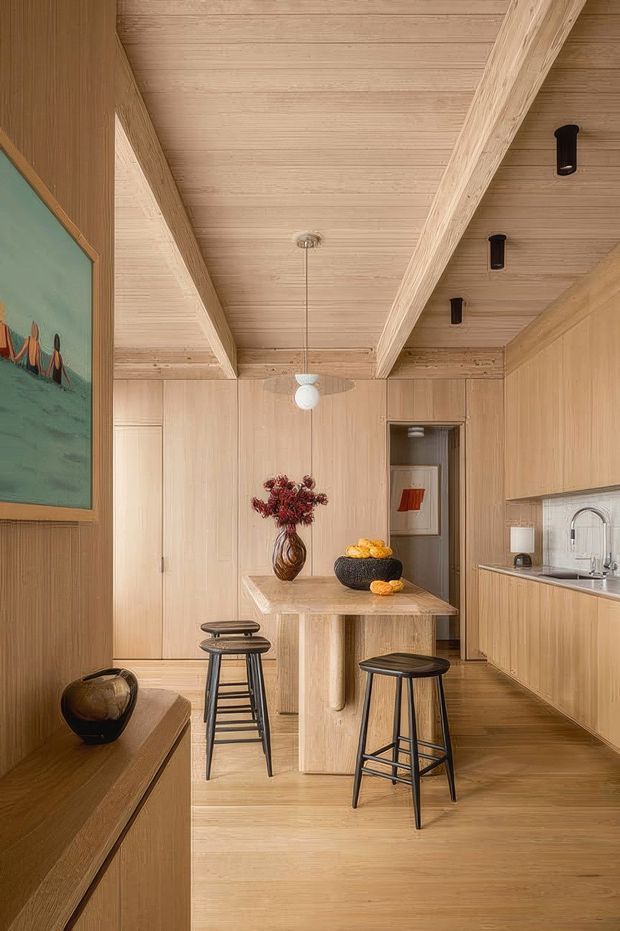 Warm, compact timber-lined kitchen with full-height light wood cabinetry, matching wood flooring and a slatted timber ceiling with exposed beams. A central stone or wood-look island provides seating for three on dark stools, while a framed artwork, pendant light, black track-style downlights, vase and bowl add restrained decorative accents. A sink and arched faucet are visible along the right-hand benchtop, with a doorway leading to an adjacent space.