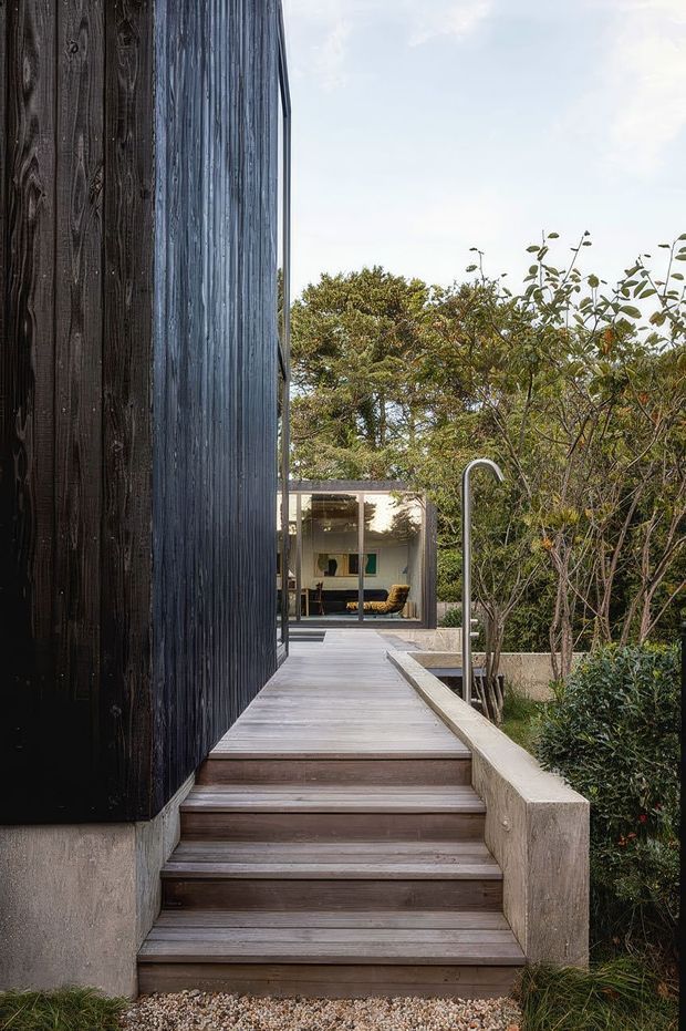 A landscaped exterior view of a modern home, showing a raised timber deck and stairway running alongside a tall dark timber-clad wall. A slim outdoor shower is positioned beside the deck, with planted garden beds and a glazed living area visible at the end of the walkway.