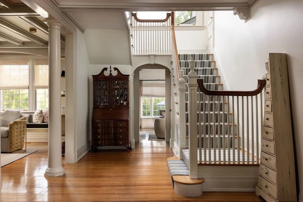 A bright, refined traditional home interior showing a polished timber hallway with substantial decorative trim, columns, and moulded ceiling details. A central staircase has white balusters, dark timber handrails, and patterned carpeted treads, while an antique dark timber display cabinet and tall chest of drawers add classic character. An adjoining sitting area is visible to the left through an open columned passage.