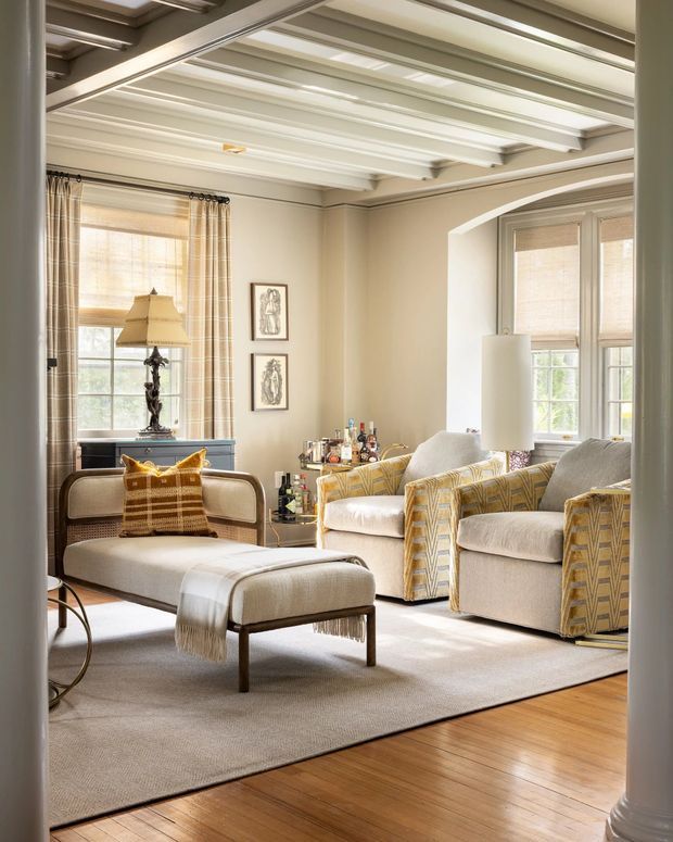 Bright, refined living room with cream upholstered seating, patterned armchairs, a chaise-style bench, and a small drinks bar. The room features warm timber flooring, a large neutral rug, tall windows with woven blinds and curtains, decorative wall art, and a distinctive white painted coffered/beamed ceiling with a broad arched opening.