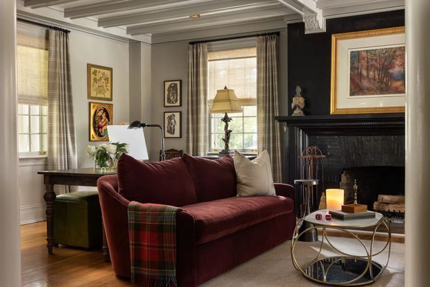 A refined traditional living room with a deep burgundy upholstered sofa, patterned throw, layered artwork, tall windows with plaid curtains, and a black carved fireplace mantel. The room features warm timber flooring, a coffered and beam-detailed ceiling, classic furnishings, and a round glass-and-metal coffee table.