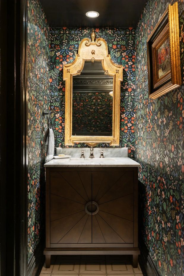 A compact, dramatic powder room with dark floral botanical wallpaper covering the walls, a black ceiling, and a dark timber vanity with radial geometric panel detailing. The vanity has a white stone or marble benchtop, polished metal tapware, and an ornate antique-style gilded mirror above it. Framed artwork and a towel rail are visible at the sides, while the floor has pale geometric patterned tiles.