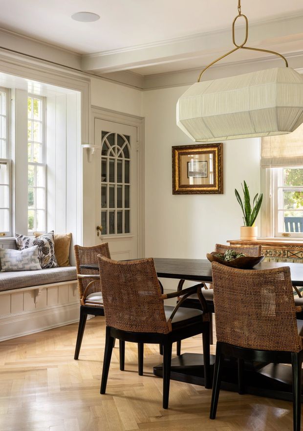 Bright, refined dining room with a large dark wood dining table surrounded by woven cane-back chairs. The room features warm painted trim and panelled cabinetry beneath a built-in window seat, tall divided-light windows, a decorative glass-panel door, a sculptural pleated pendant light, framed artwork, and a potted plant. Light herringbone timber flooring and abundant daylight create an inviting, elegant atmosphere.