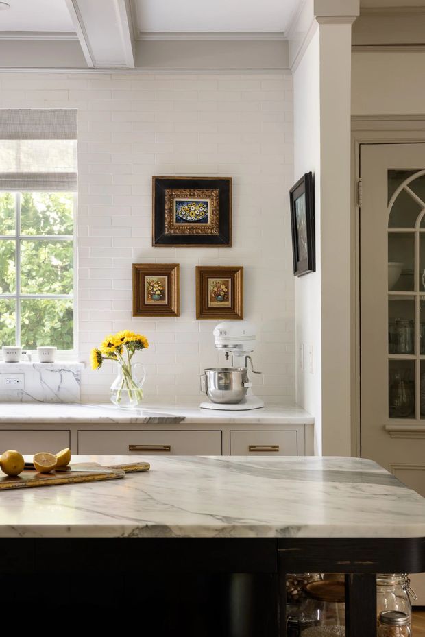 Bright, traditionally styled kitchen with a large white marble island in the foreground, dark timber cabinetry below, and a pale marble countertop along the rear wall. The room features white-painted brick walls, decorative ceiling mouldings, a large divided-pane window with a Roman blind, framed artwork, brass-toned cabinet pulls, and a glass-fronted built-in cupboard. A stand mixer, fruit, and flowers add lived-in detail.