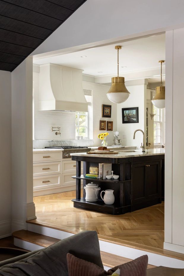 Bright, refined kitchen viewed from an adjacent living area, featuring a large dark island with open display shelving, pale cream cabinetry, a marble-look benchtop, white tiled splashback, herringbone timber flooring, brass pendant lights, and a sculptural rangehood. A small portion of the living room is visible in the foreground.