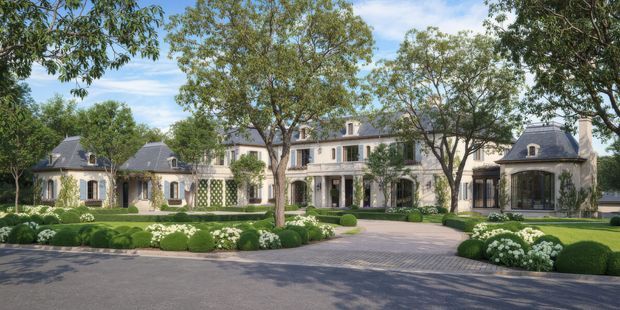 A grand symmetrical residential estate viewed from the street, featuring a long pale masonry facade with multiple connected wings, steep dark slate-like hipped roofs, dormer windows, classical columns, tall glazed openings, and manicured formal gardens with clipped hedges and rounded shrubs. A broad paved driveway leads to the central entrance, giving the property a refined European-inspired, estate-like character.