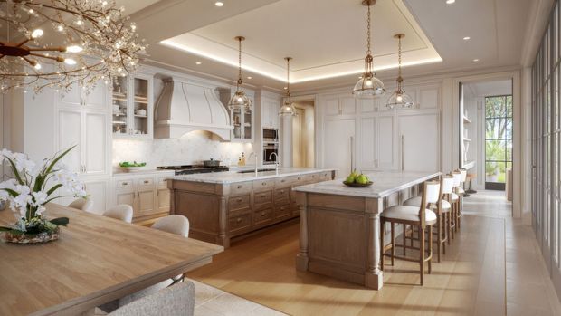 A spacious, refined traditional kitchen with two large timber-topped islands, extensive white raised-panel cabinetry, a marble-look splashback, integrated appliances, and multiple pendant lights. A dining table occupies the foreground, while a row of bar stools lines the main island. Tall glazed doors and windows bring in daylight and overlook greenery.