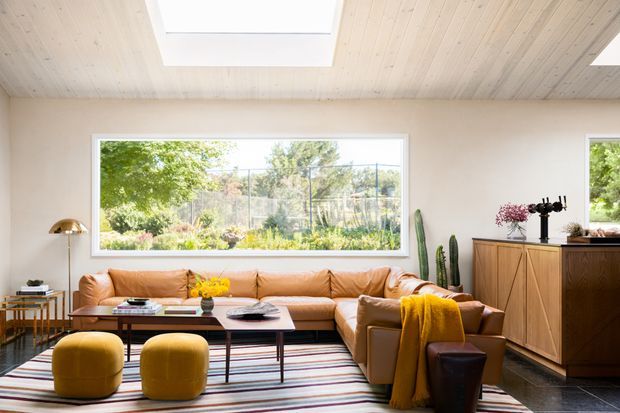 Bright contemporary living room with a low tan leather sectional sofa, mustard ottomans, a striped area rug, dark timber coffee tables, and a timber sideboard. The room features a pale timber-lined sloping ceiling with a large skylight, white walls, expansive garden-facing glazing, and indoor plants. Warm natural light creates an inviting, relaxed atmosphere.