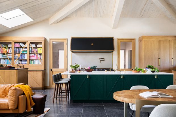 Bright open-plan kitchen and dining area with a dark green island, timber cabinetry and bookcase joinery, black tiled flooring, and a vaulted light timber ceiling with exposed beams and skylights. A lounge chair and sofa edge are visible in the foreground, while a round timber dining table occupies the right side.