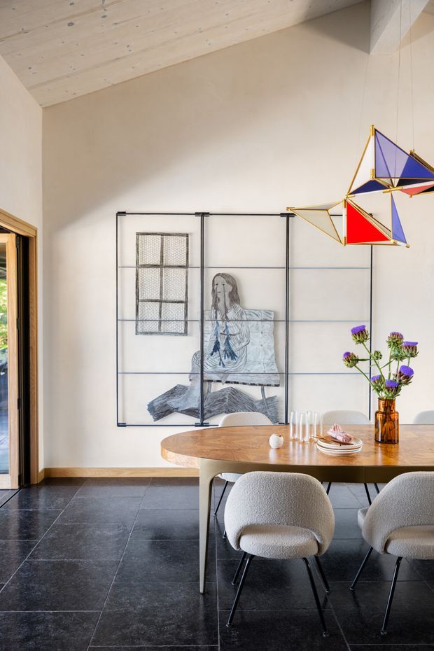 A refined dining area with a large oval timber dining table and upholstered cream boucle chairs on dark stone tile flooring. A black metal-framed artwork or room divider with a monochrome seated figure occupies the wall, while a colourful geometric pendant light hangs above the table. The room has warm plaster walls, a pale timber-lined sloping ceiling, and a timber-framed glazed opening at left.