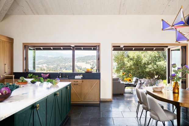 Bright, spacious open-plan kitchen and dining area with a large dark green island, white benchtop, timber cabinetry, and dark tiled flooring. Expansive glazed openings connect the dining area to a leafy outdoor setting and distant hills. A sculptural geometric pendant hangs above the dining table, adding a colourful decorative accent. The interior combines natural timber, greenery, and abundant daylight.