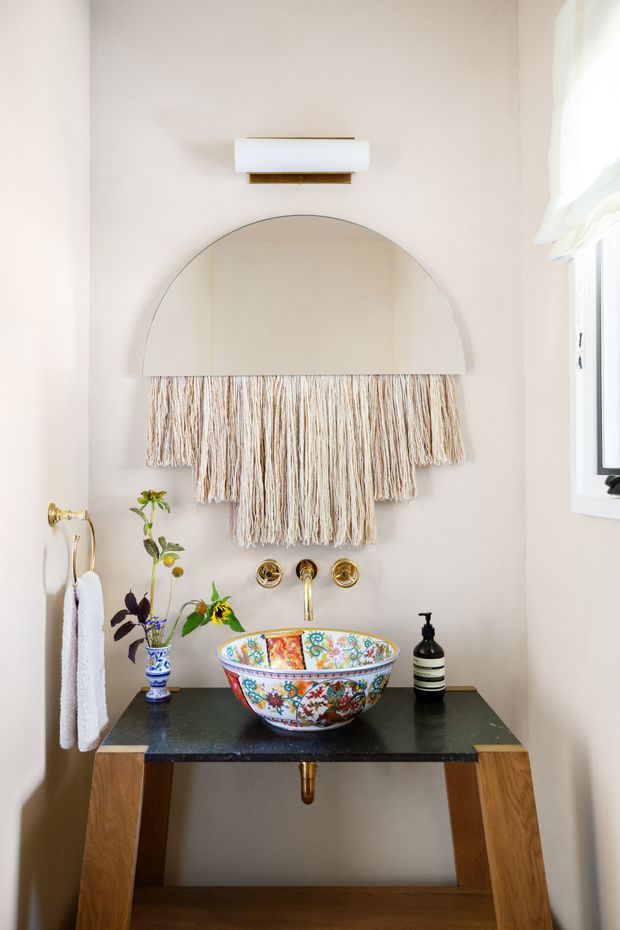 A compact, elegantly styled bathroom vanity with a freestanding patterned ceramic vessel basin on a dark stone benchtop and timber console base. Brushed brass wall-mounted tapware and a brass-framed arched mirror sit above the vanity, with a textured woven wall hanging beneath the mirror. A small floral arrangement, soap dispenser, and hand towels add decorative detail. The space is softly lit and finished in warm off-white tones with a black-framed window at right.