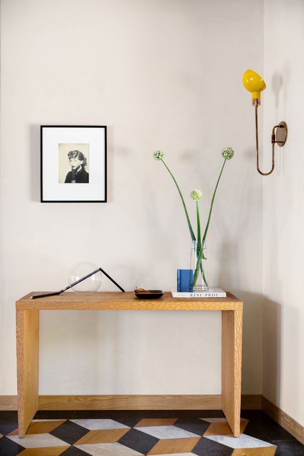 A styled entryway vignette with a simple natural timber console table against a warm off-white wall. The console displays a framed black-and-white portrait, modern table lamp, small dish, blue book, and tall glass vase with delicate green stems. A sculptural brass-and-yellow wall sconce is mounted above, while the floor features a bold geometric pattern of timber, black, and pale stone-like tiles. The composition has a refined mid-century-influenced contemporary feel.