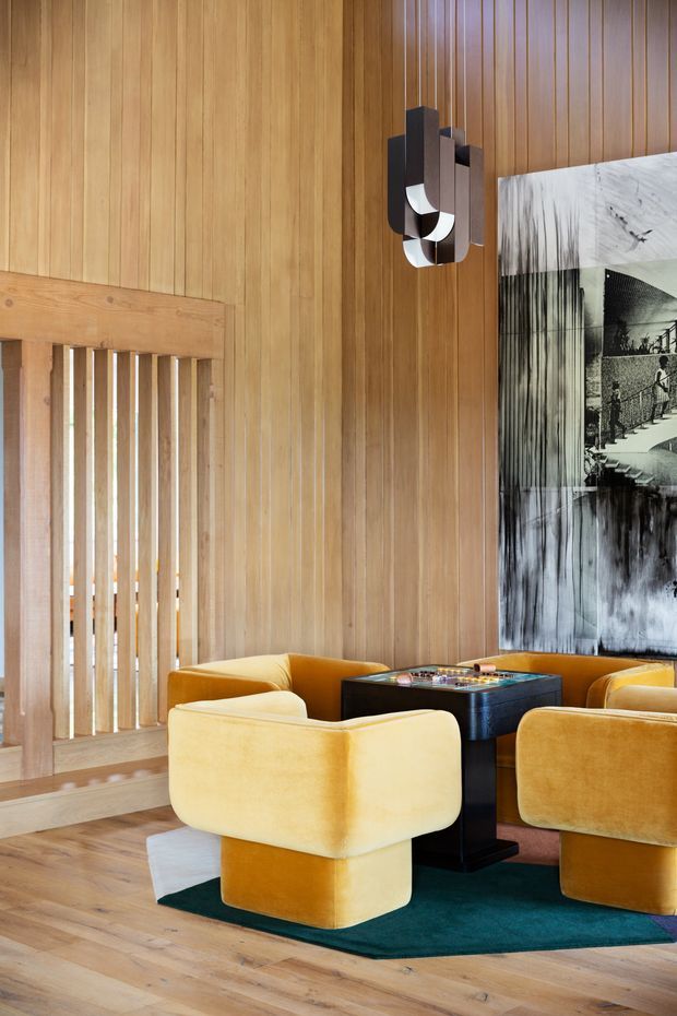 A warm, richly detailed lounge area with vertical timber wall panelling, timber flooring, and a timber slatted divider. Several rounded mustard-yellow upholstered lounge chairs surround a dark square table, set on a deep teal rug. A sculptural wall light and a large monochrome artwork provide dramatic accents. The space has a refined mid-century-inspired character with strong timber detailing and bold furniture forms.