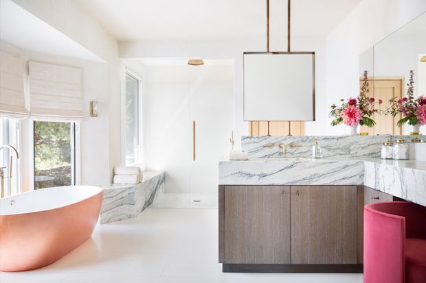 Bright, luxurious contemporary bathroom with a freestanding copper-toned bathtub, a marble-clad vanity island, integrated basin, large suspended rectangular mirror, and warm timber cabinetry. A shower or wet area is visible at the rear, while a vivid pink upholstered stool adds a bold accent. The room has abundant natural light and a clean, refined palette of white, marble grey, timber, copper, and pink.